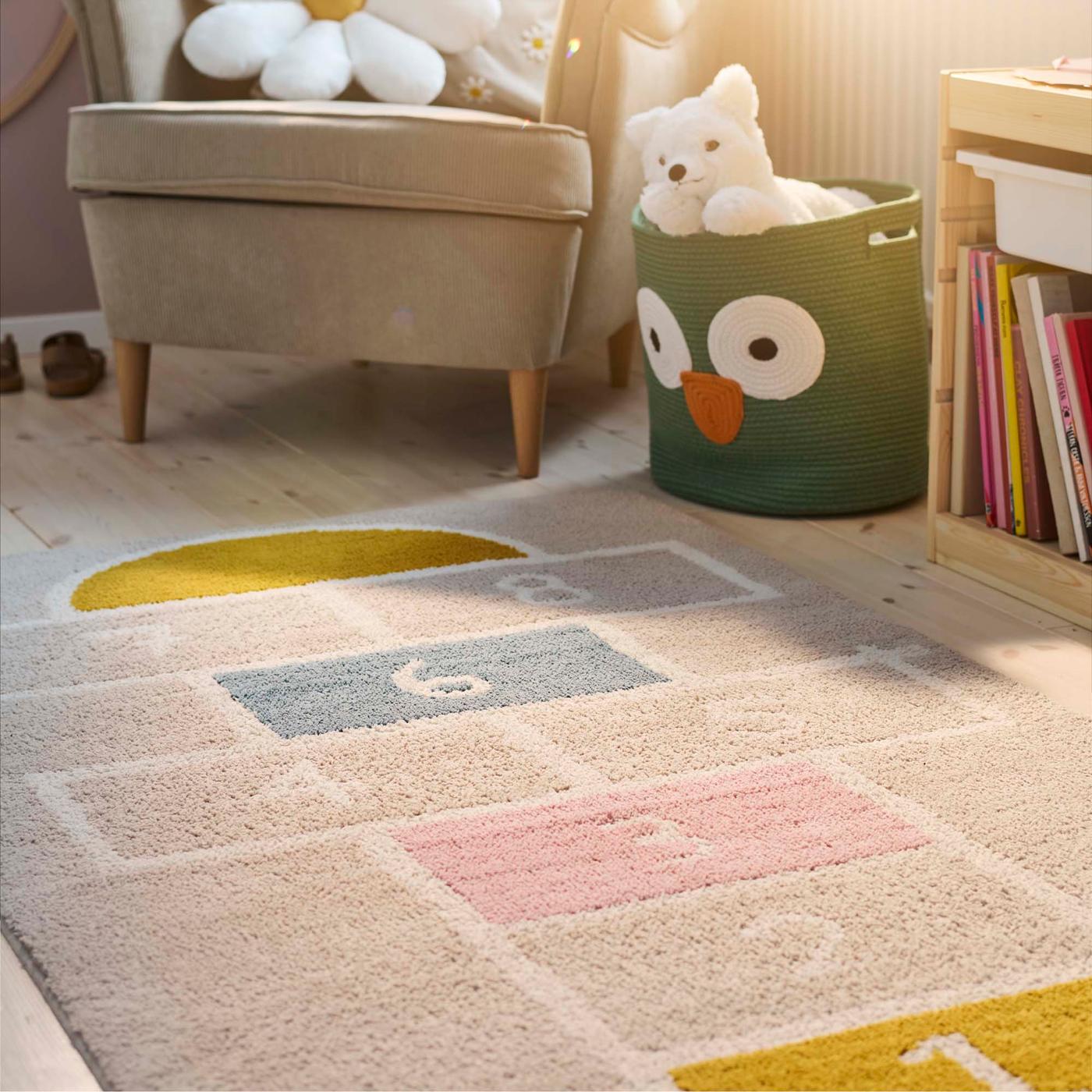 A toy bin has a motif of an owl's face with two big eyes and a beak. A white stuffed toy bear peeks out of the top of the bin. Part of a rug with a hopscotch pattern is in the foreground.