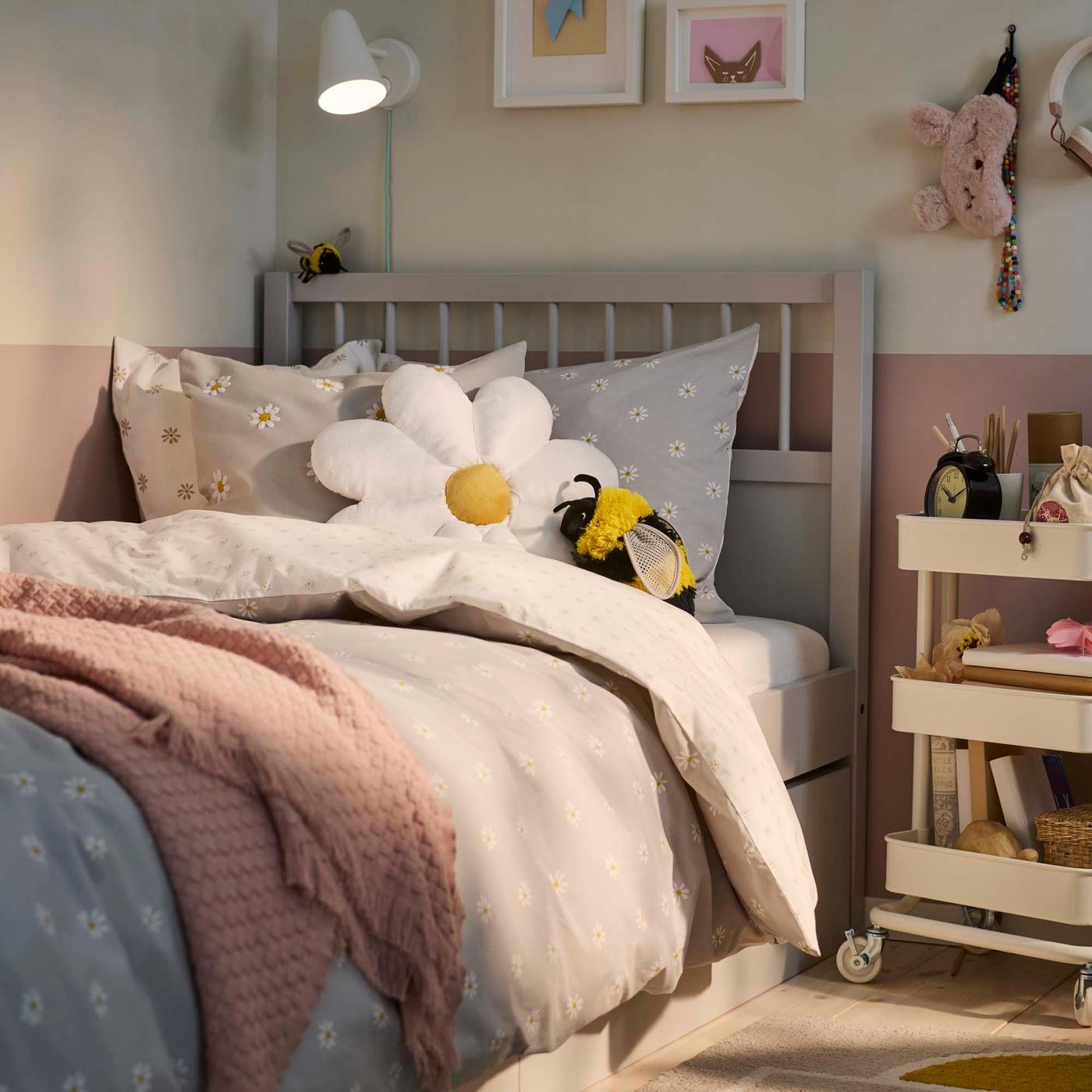 A bed is dressed with a pale grey duvet cover and a dusty pink blanket. Two soft toy cushions are on the bed, in the shapes of a daisy and a bumblebee.