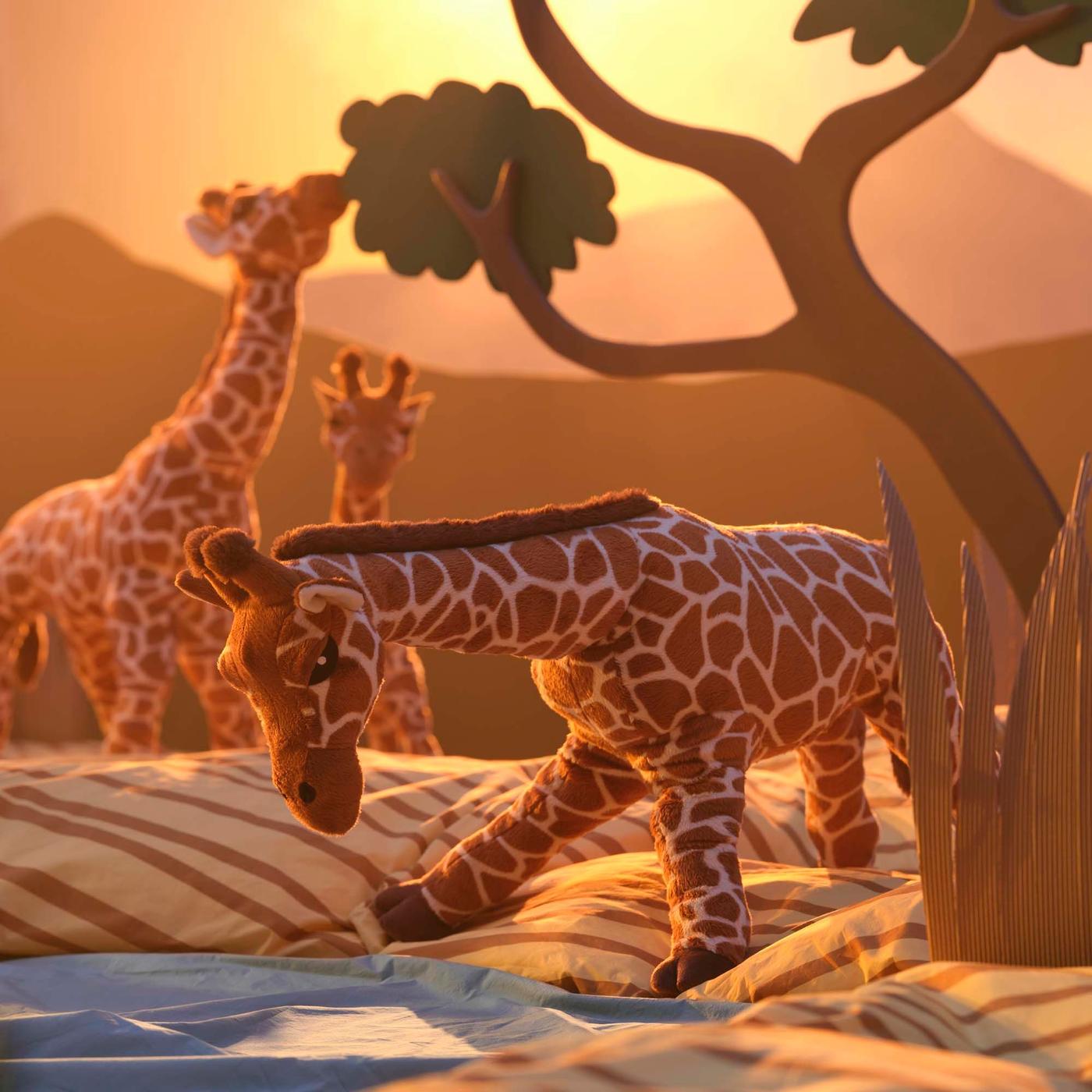 Three soft toy giraffes beside a tree and a pond in an imaginary setting of the African savannah.