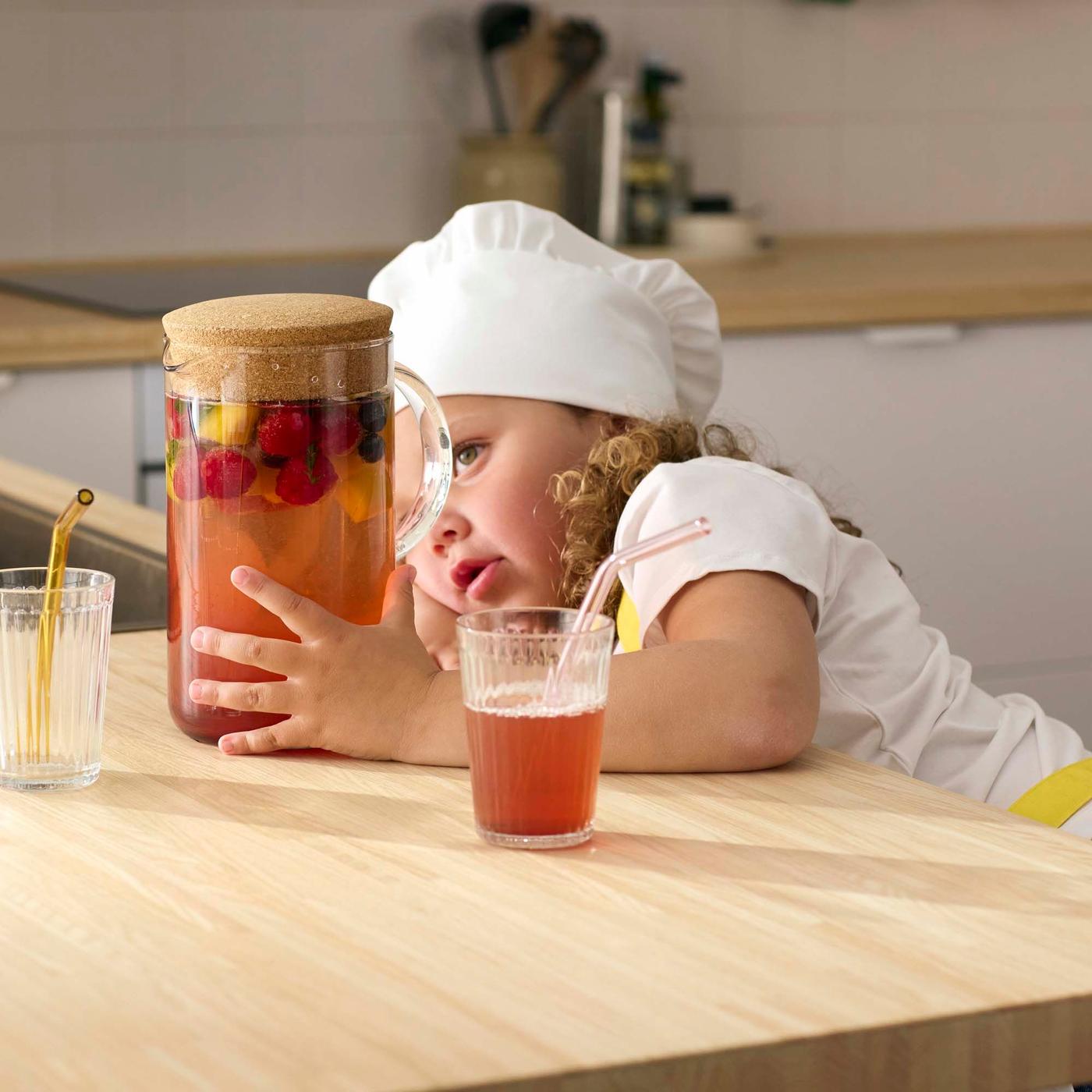 A young child in a white and wood kitchen wearing a chefs hat with a glass jug filled with juice and fresh fruit. 