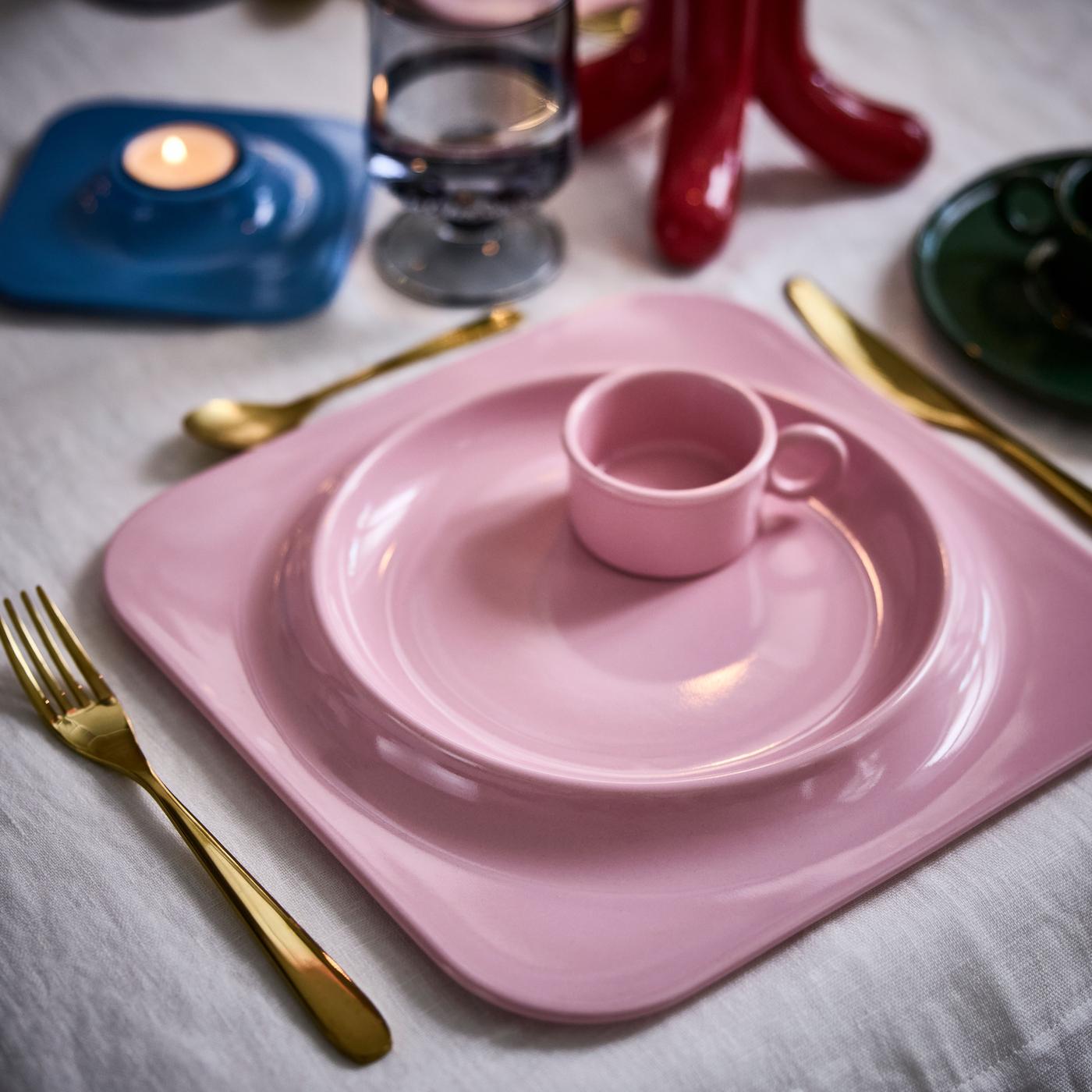Pink square porcelain plate seen from above with a pink cup inside, lit candles and gold cutlery beside it.