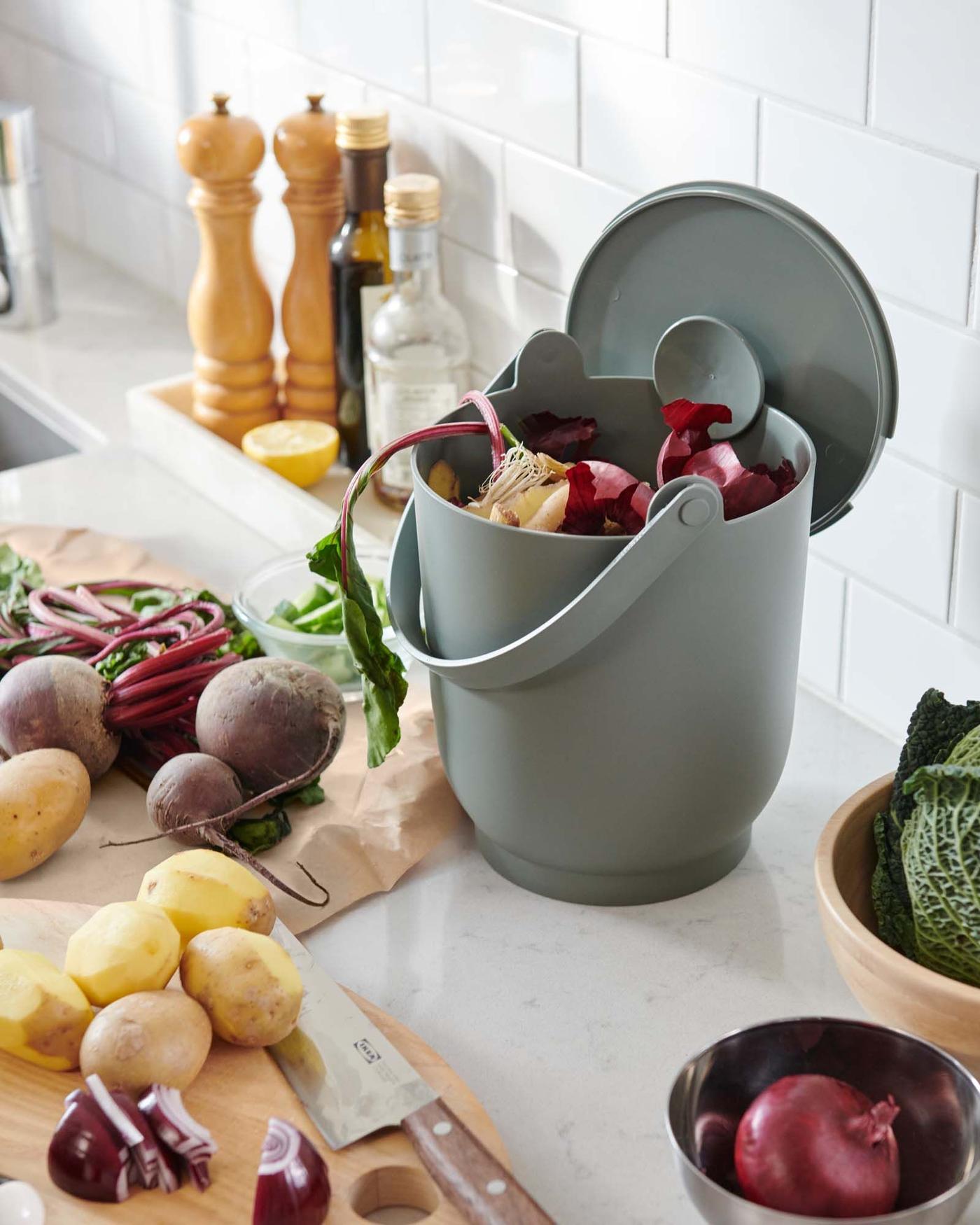 A grey-green FARMARKVAST bin filled with food waste sits on a kitchen counter, surrounded by vegetables.