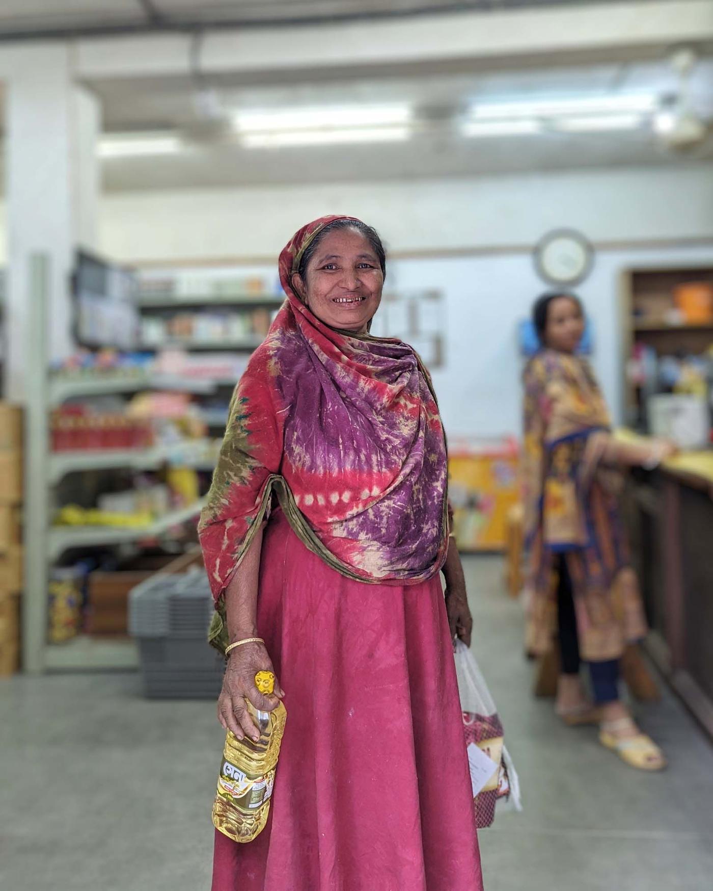 A woman dressed in pink stands in a grocery shop. She smiles as she holds groceries in her hands. A lady stands behind her in the distance.