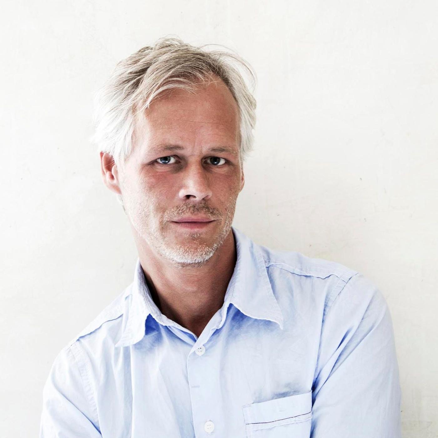 A portrait of Hans Blomquist wearing a light blue shirt and standing against a white wall.