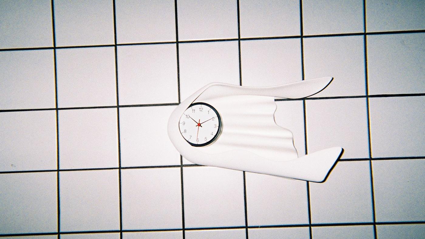 A white wall clock with a plastic frame resembling a flowing curtain, on a white-tiled wall.