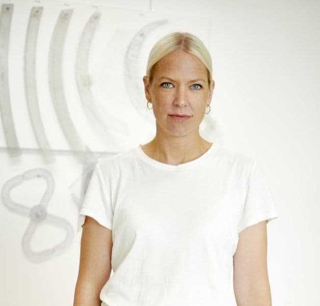 A woman in a white t-shirt stands in front of a white wall with patterns on it.