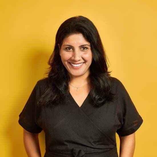 A woman wearing black stands against a yellow background. She is smiling.