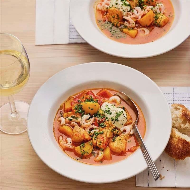 Spicy salmon stew with shrimp and aioli served in a white bowl atop a light wood table.