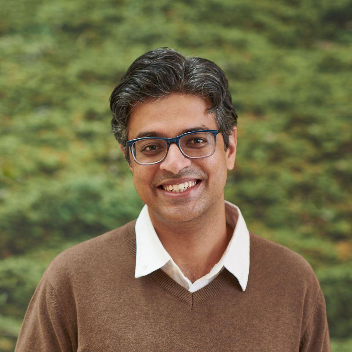 Portrait of Sriram Rajagopal wearing a white shirt, brown pullover and standing against a wall of greenery.