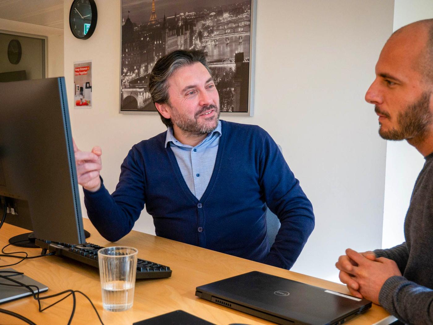 Two people sit at a desk with a computer. One looks at the screen while the other speaks.