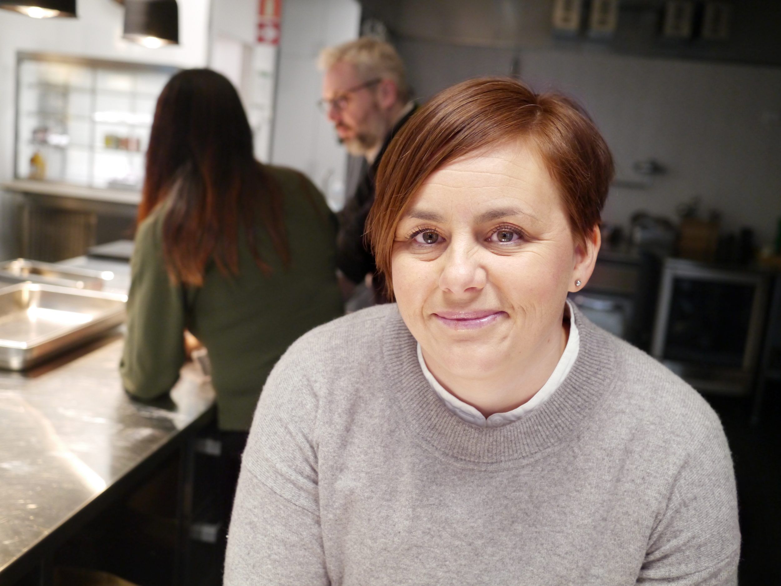 A smiling woman standing in a professional kitchen with two people in the background.