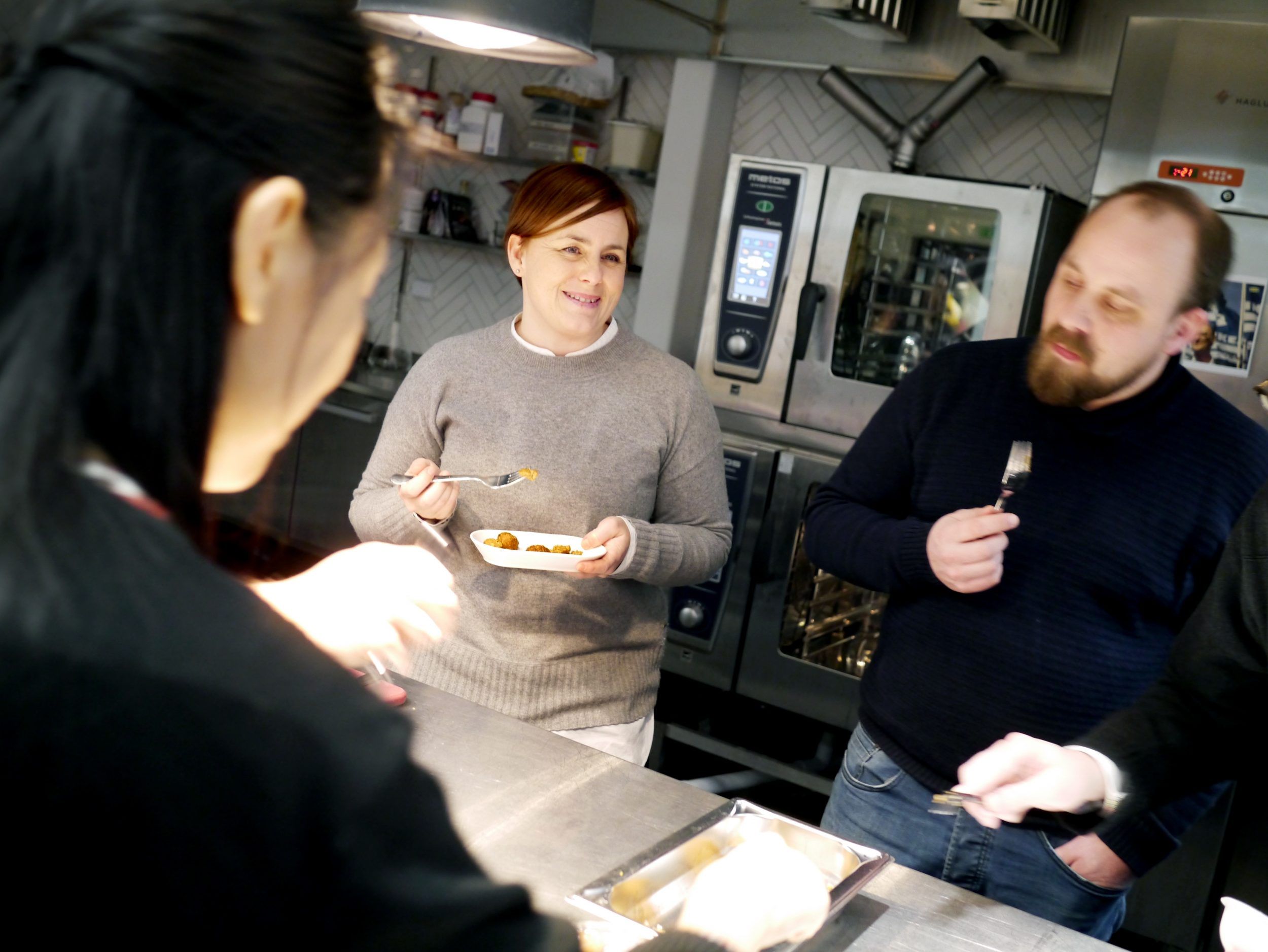 Three people standing in a professional kitchen, eating plant balls.