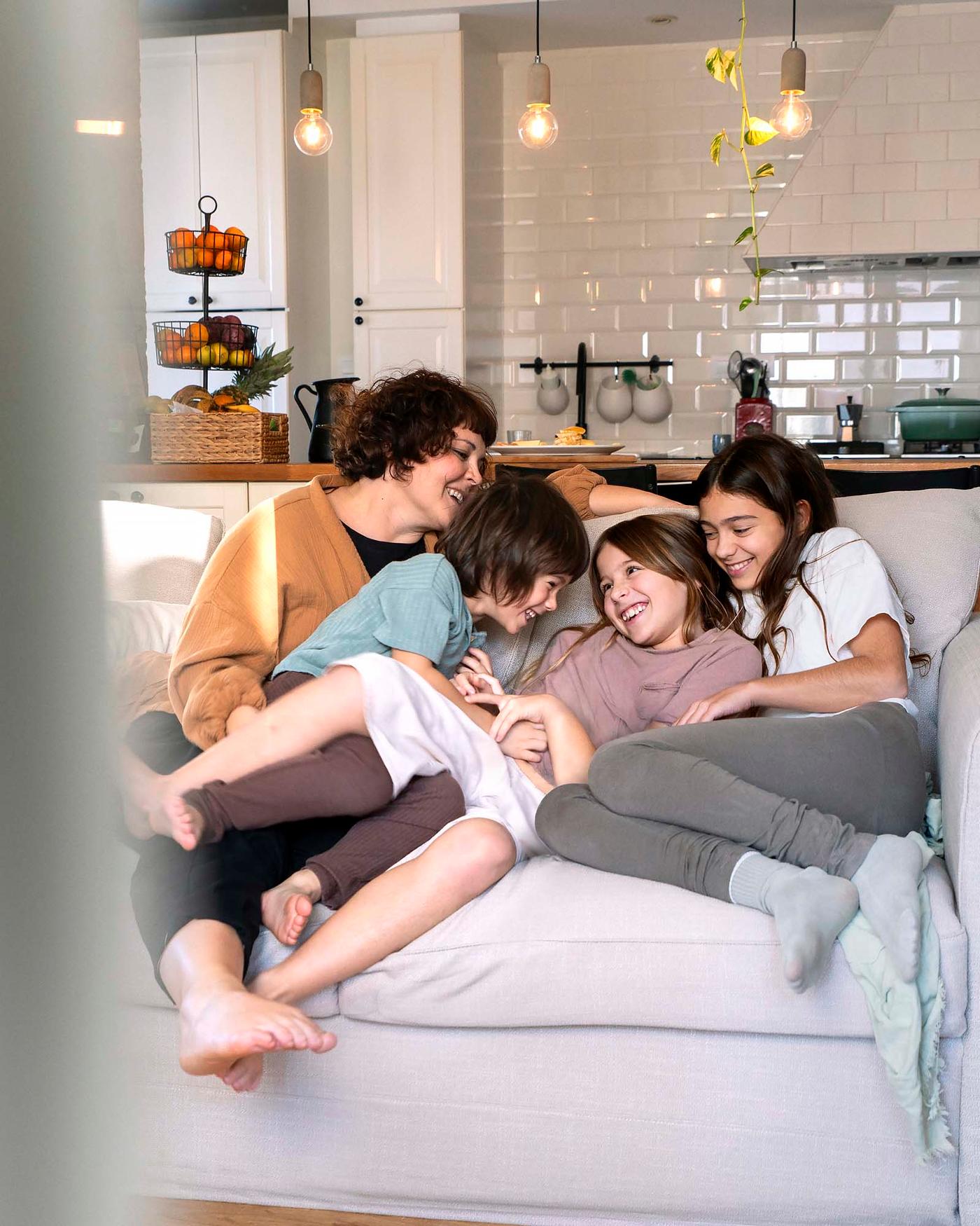 An adult and three children laugh as they sit closely together on a sofa. There is a kitchen area behind.