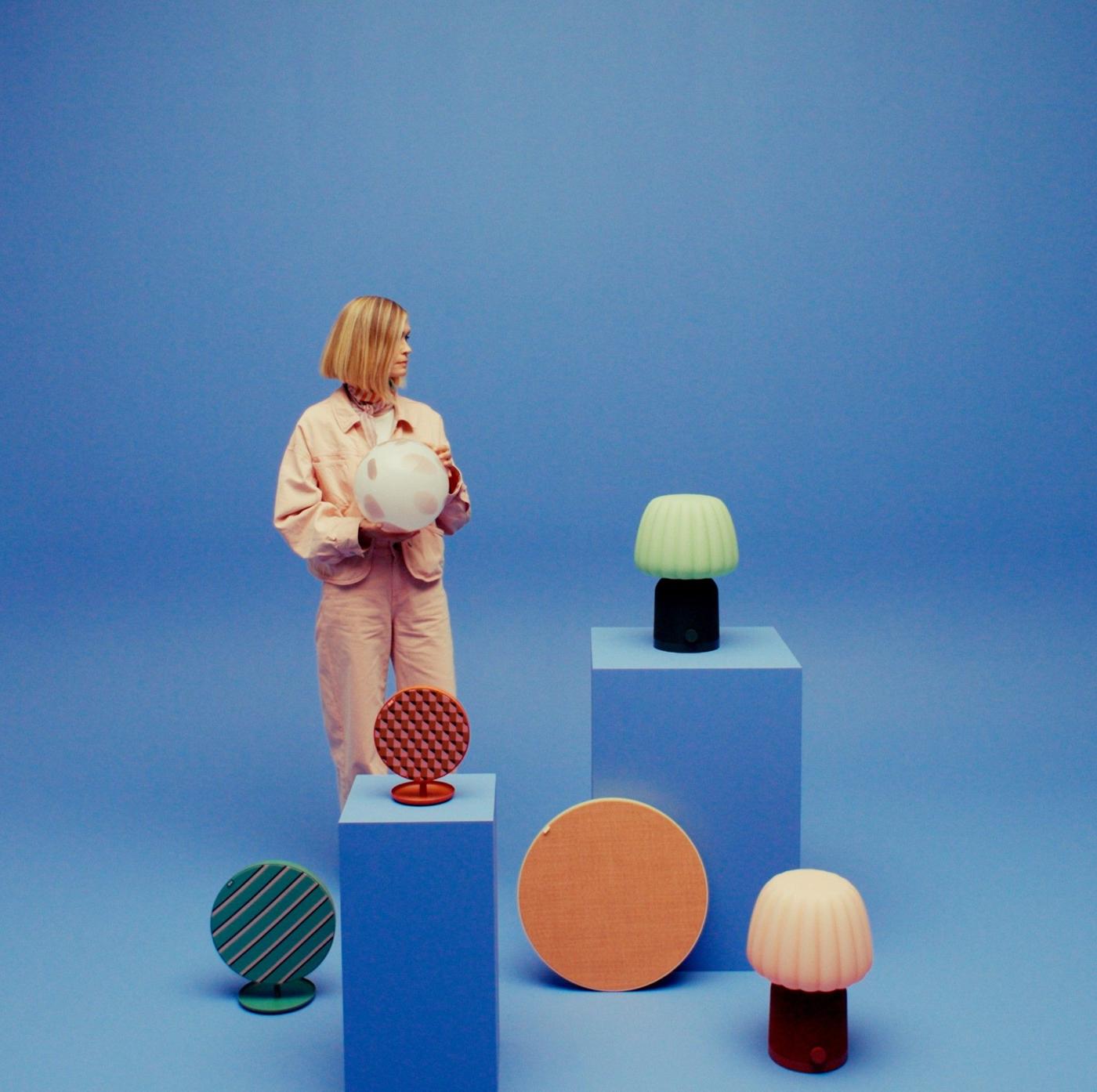 Tekla Severin stands among various speakers placed on pedestals, holding a spherical spotted lamp against a blue backdrop.