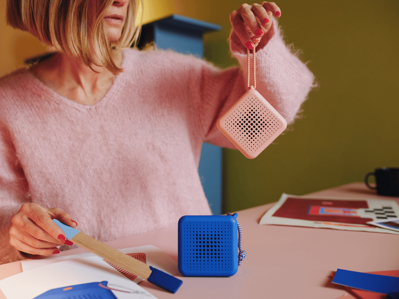 Tekla Evelina Severin is holding up a pink portable bluetooth speaker in its leash while the blue bluetooth speaker stands next to it. She's holding a wooden stick that has been dripped in colour.