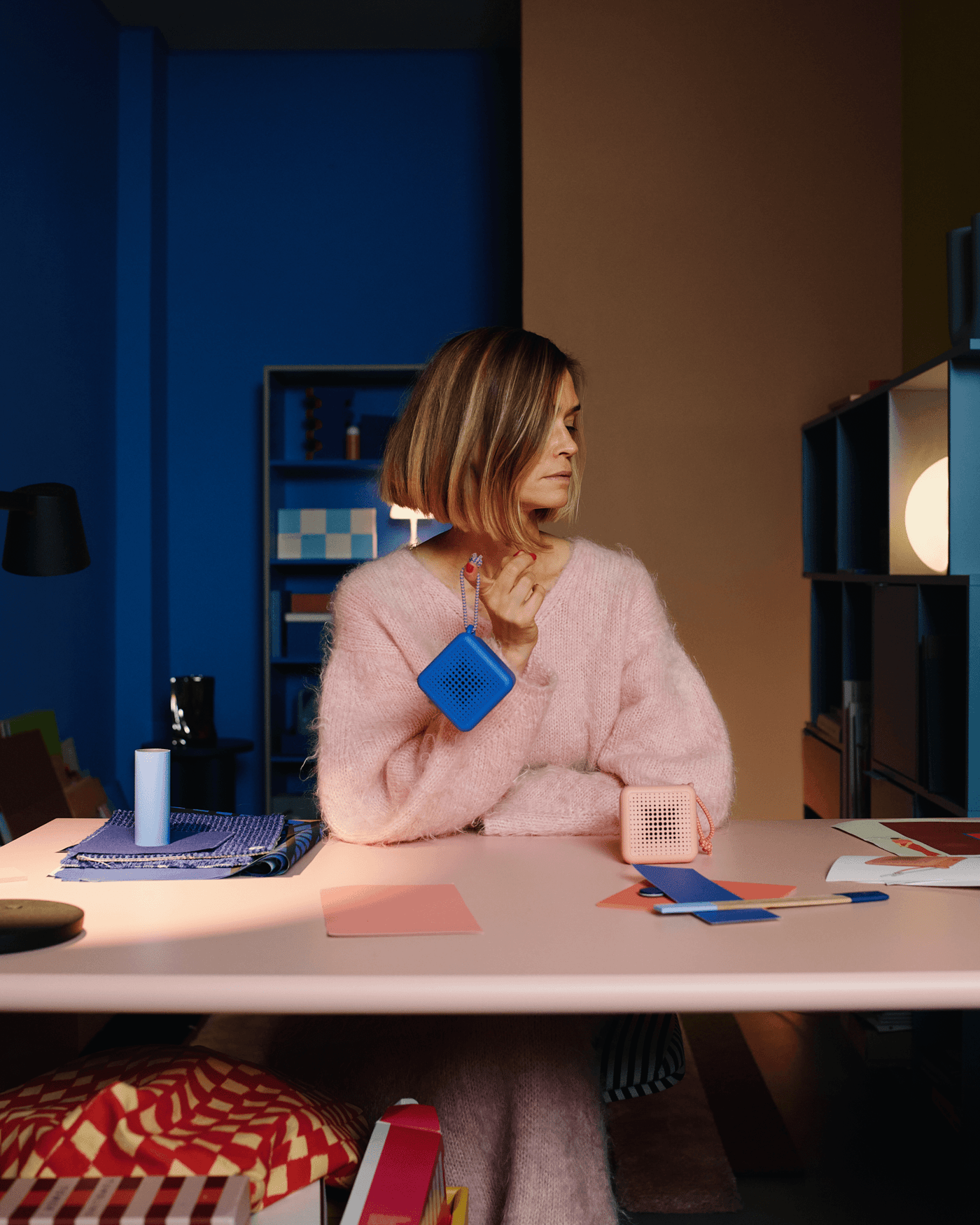 Multidisciplinary designer and colourist Teklan sits at desk, in pink and turned away, holding a blue portable speaker in her right hand, with a pink speaker to her left.