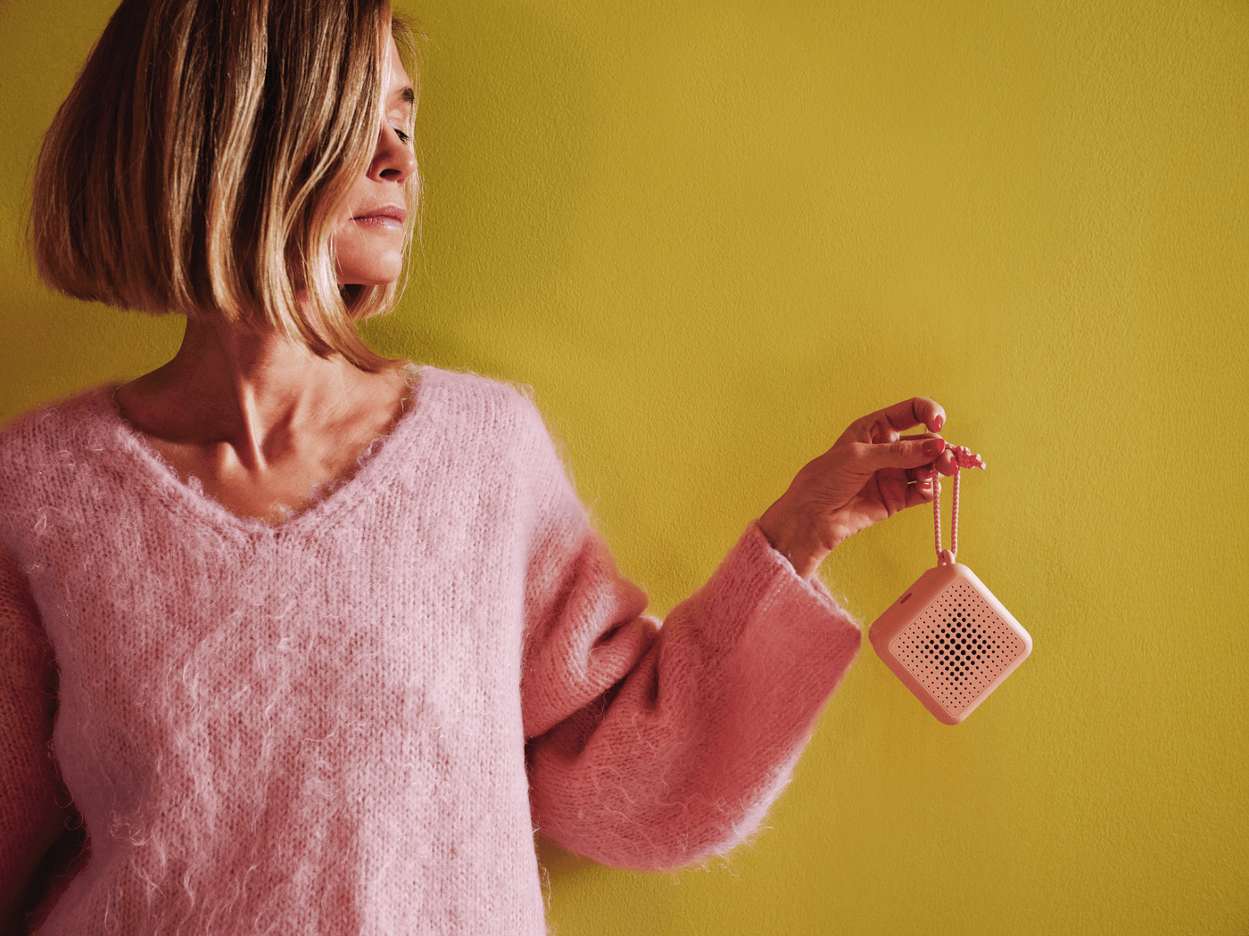 Tekla Evelina Severin is dressed in pink standing against a yellow wall holding up a small pink portable VAPPEBY speaker, holding it's leash.