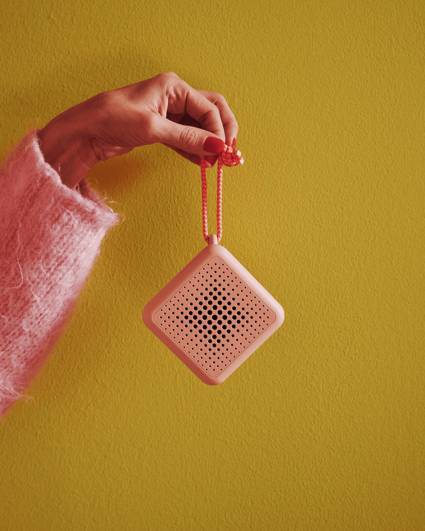 A hand is holding a pink portable speaker in its leash. The background wall is yellow.