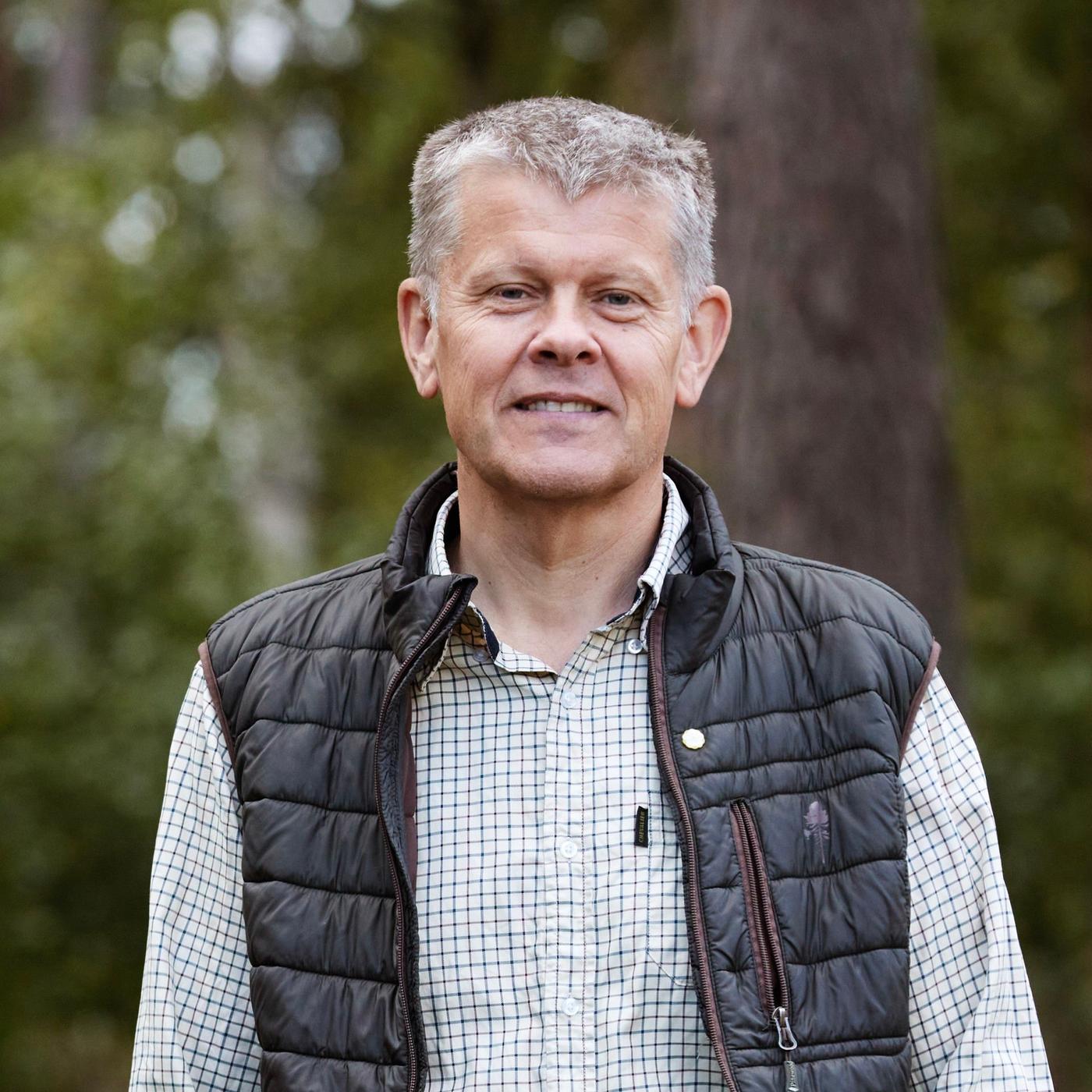 A portrait of a man in a checkered shirt and a vest standing in the forest.