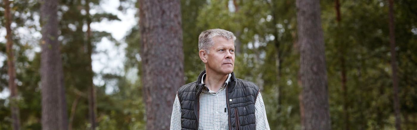 A man in a checkered shirt and a vest standing in the forest.