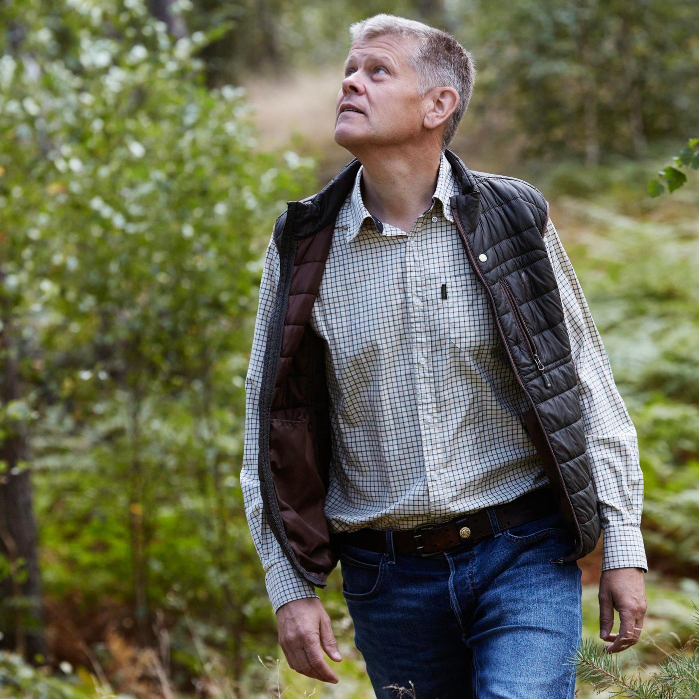 A man in a checkered shirt and a vest walking in the forest.