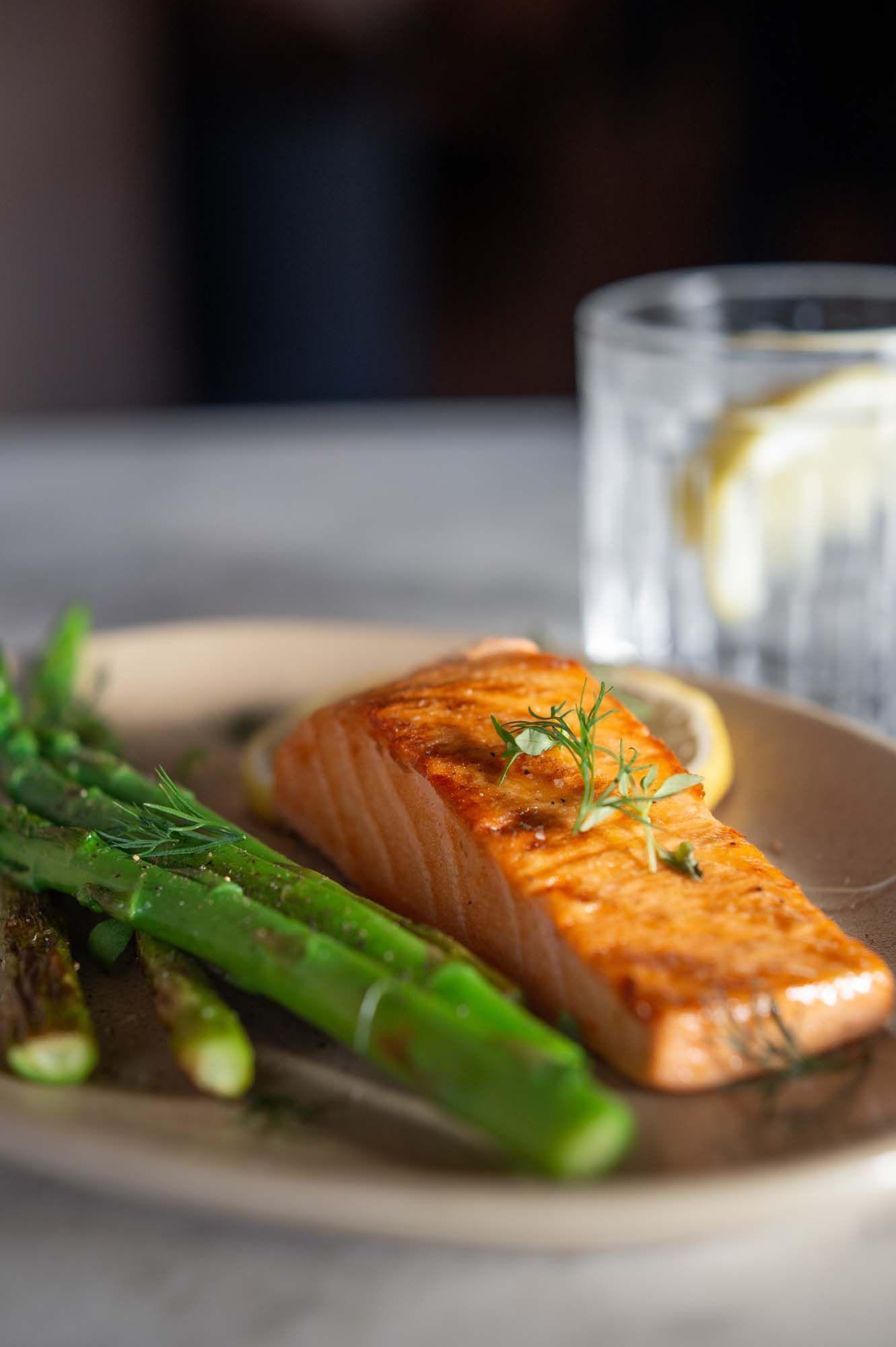 A piece of cooked, New School Foods salmon sits on a plate. It has green garnish on top and green vegetables beside it. There is a glass with water and lemon in the background.