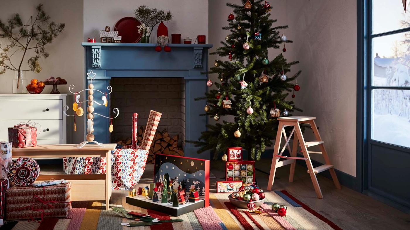 A room filled with Christmas decorations. Next to a blue fireplace stands a christmas tree being decorated. A wooden step stool stands next to it.