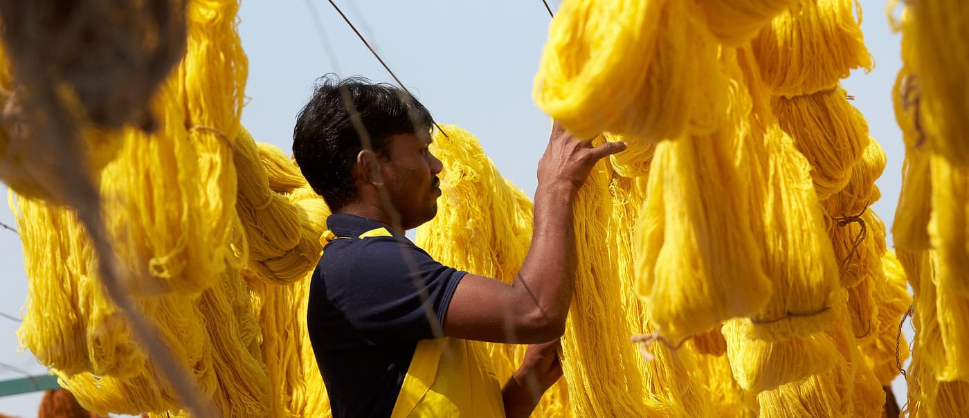 A man in a yellow apron stands in the middle of rows of yellow yarn hung out to dry in the sun.