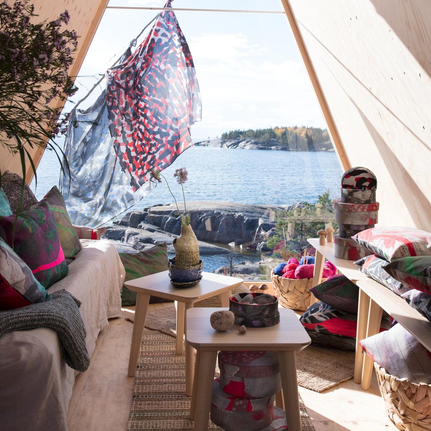 A teepee-like tent in front of a sunny lake with a sofa, chairs for tables and products from the ANNANSTANS collection.