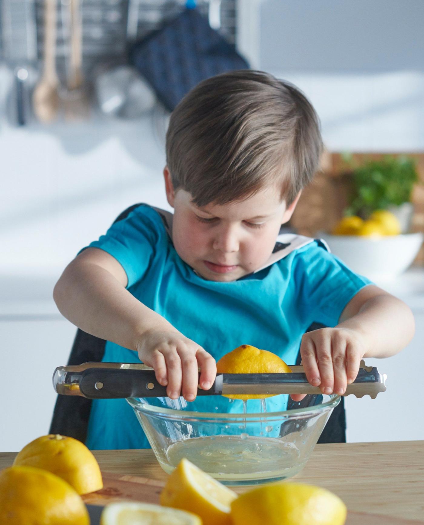 A boy in a blue shirt and a cape squeezing a lemon with VARDAGEN cooking tweezers over a VARDAGEN glass bowl.