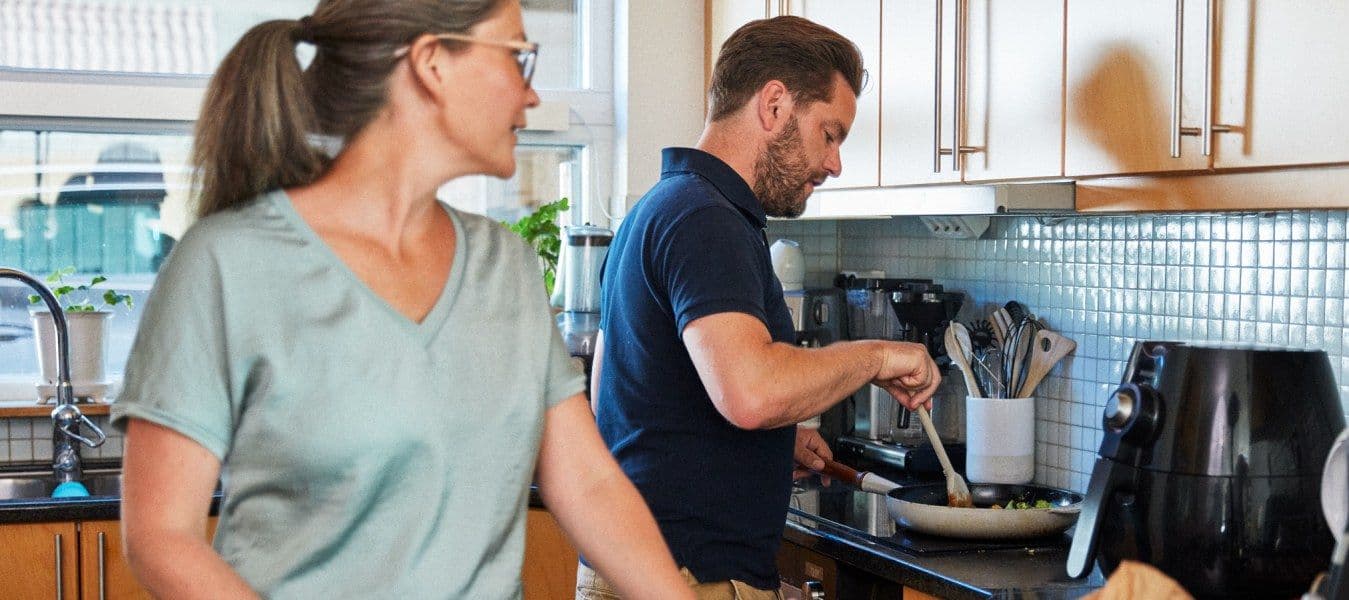 In a kitchen, a couple prepare a meal. A woman talks over her shoulder to a man, who is stirring food in a frying pan.