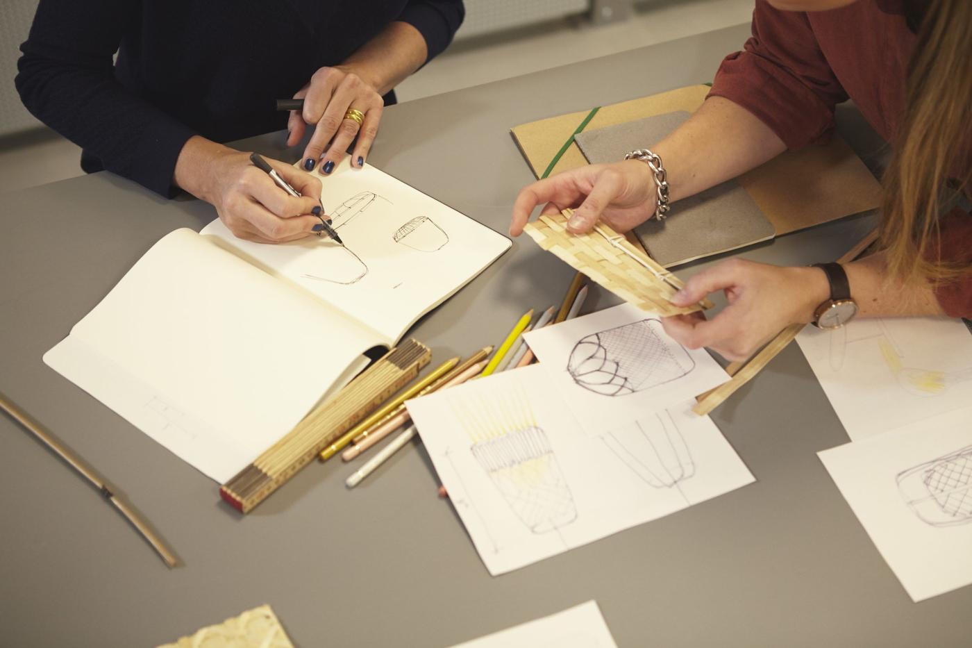 Two pairs of women's hands working with sketches and samples of woven bamboo at a grey table.