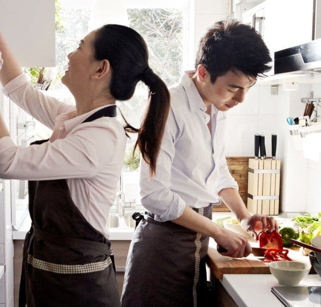 A young man and woman cooking together in a tiny, white kitchen with glass, metal, stoneware and wooden accessories.