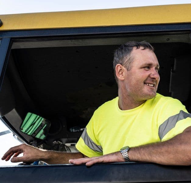 A man in a yellow T-shirt sitting in a truck with his left arm out the window looking back, backing the truck.