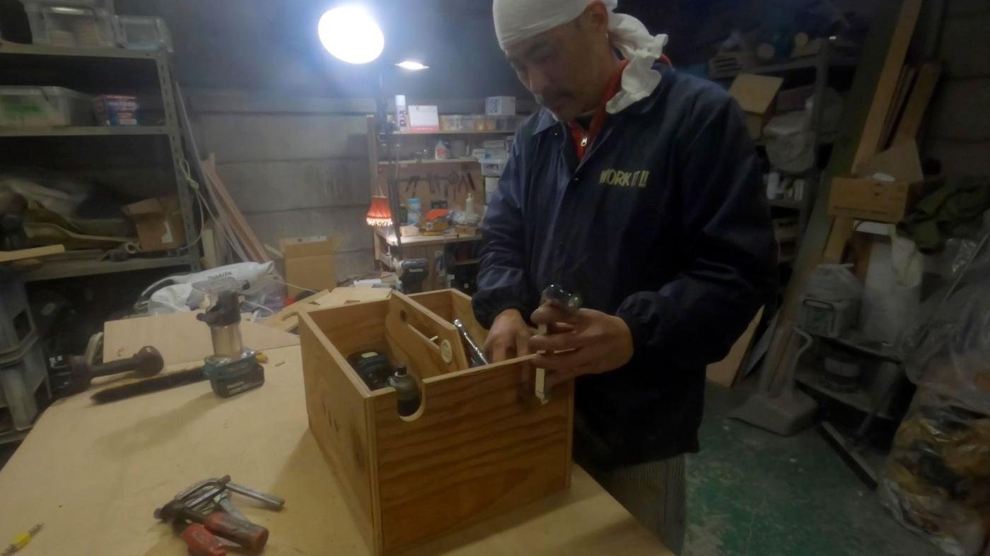 A man in a blue jacket working with a toolbox at a workbench in a workshop, with shelving in the background.