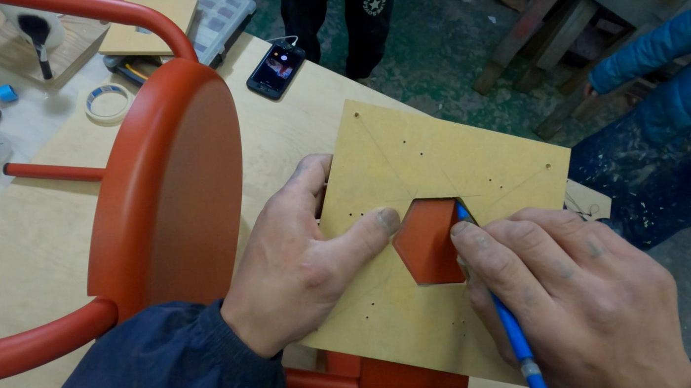 A pair of hands drawing with a hexagonal template on the backrest of a red chair on a workbench.