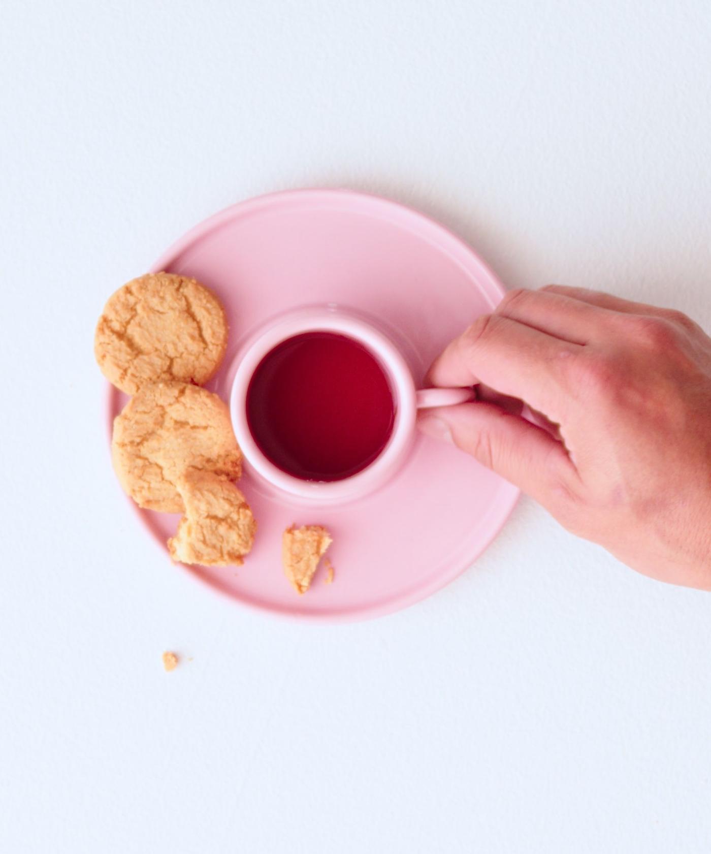 A hand holds the handle of a pink cup holding tea, viewed from a top down angle. On the pink saucer that the teacup is resting on there are three cookies. One has a bite taken from it.
