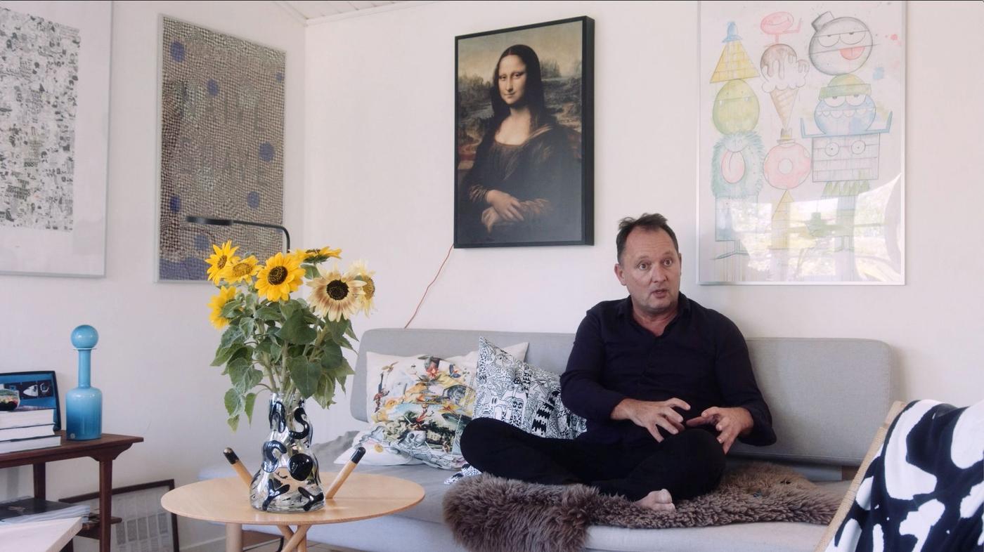Henrik Most sitting in a grey sofa with cushions, with the Mona Lisa and other framed art prints on the wall behind him.
