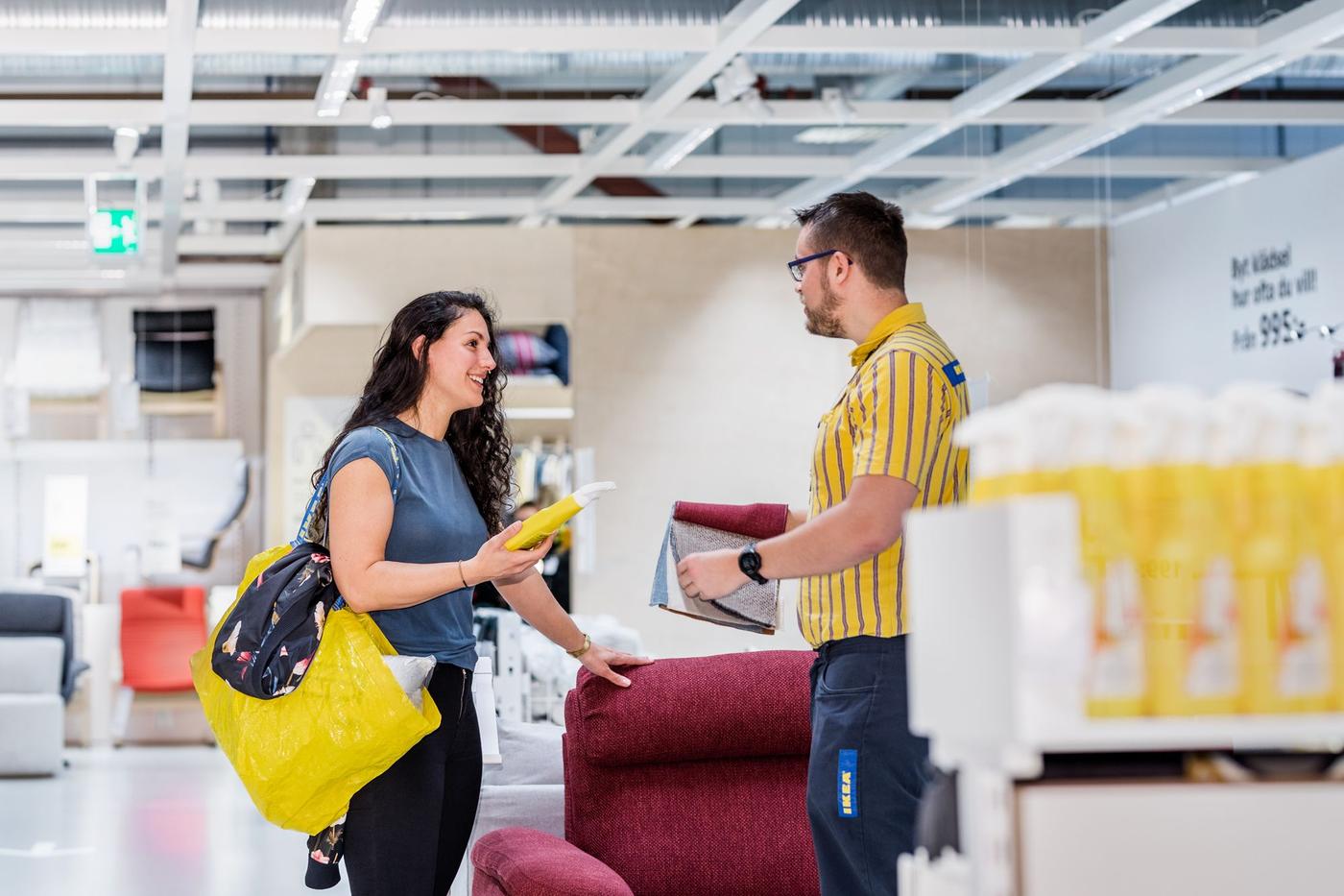 A customer wearing a blue shirt and carrying a yellow a yellow IKEA bag asks an IKEA co-worker for help in an IKEA store.