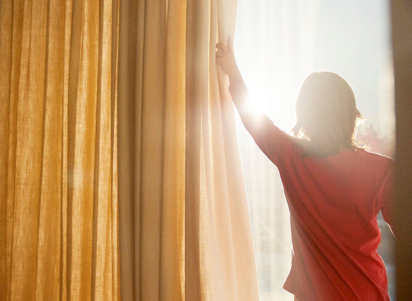 A person wearing a red t-shirt opening curtains to reveal bright sunshine streaming in.