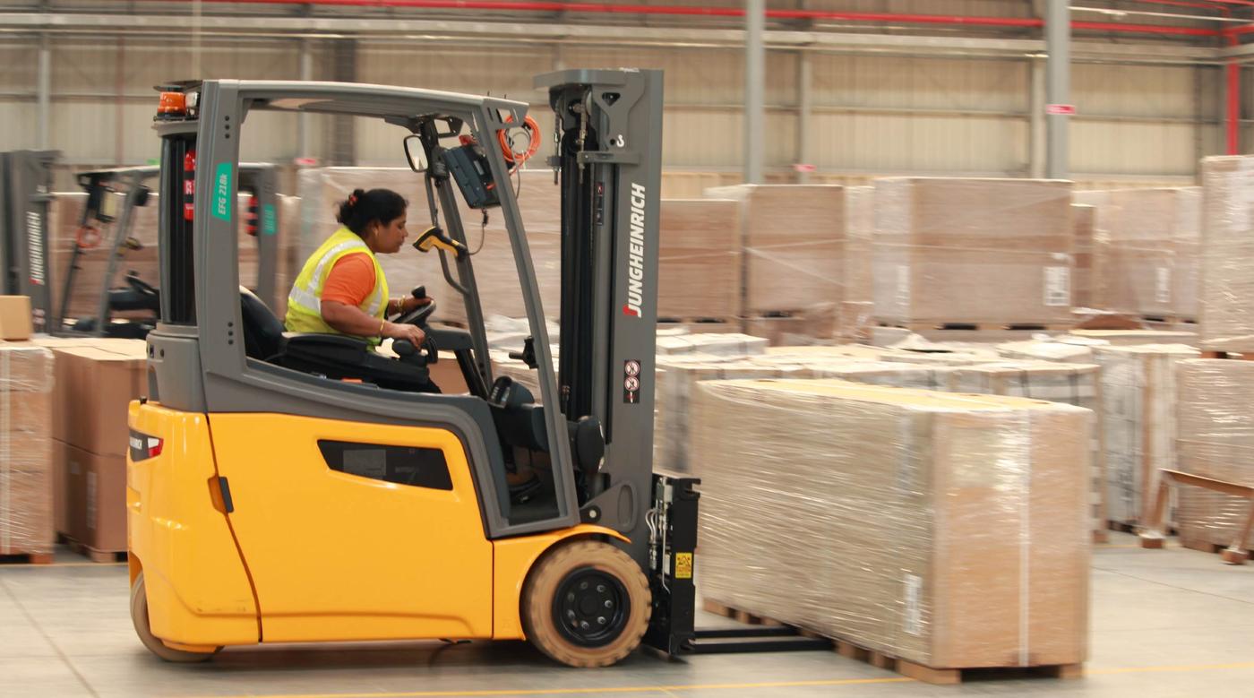 A forklift truck lifts a large wrapped box inside a warehouse. The woman driving it is wearing a yellow safety vest.