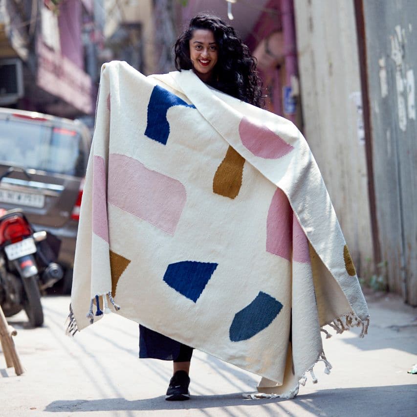 A woman on a city street, holding a white flat-woven rug with blue, brown and pink shapes on it.