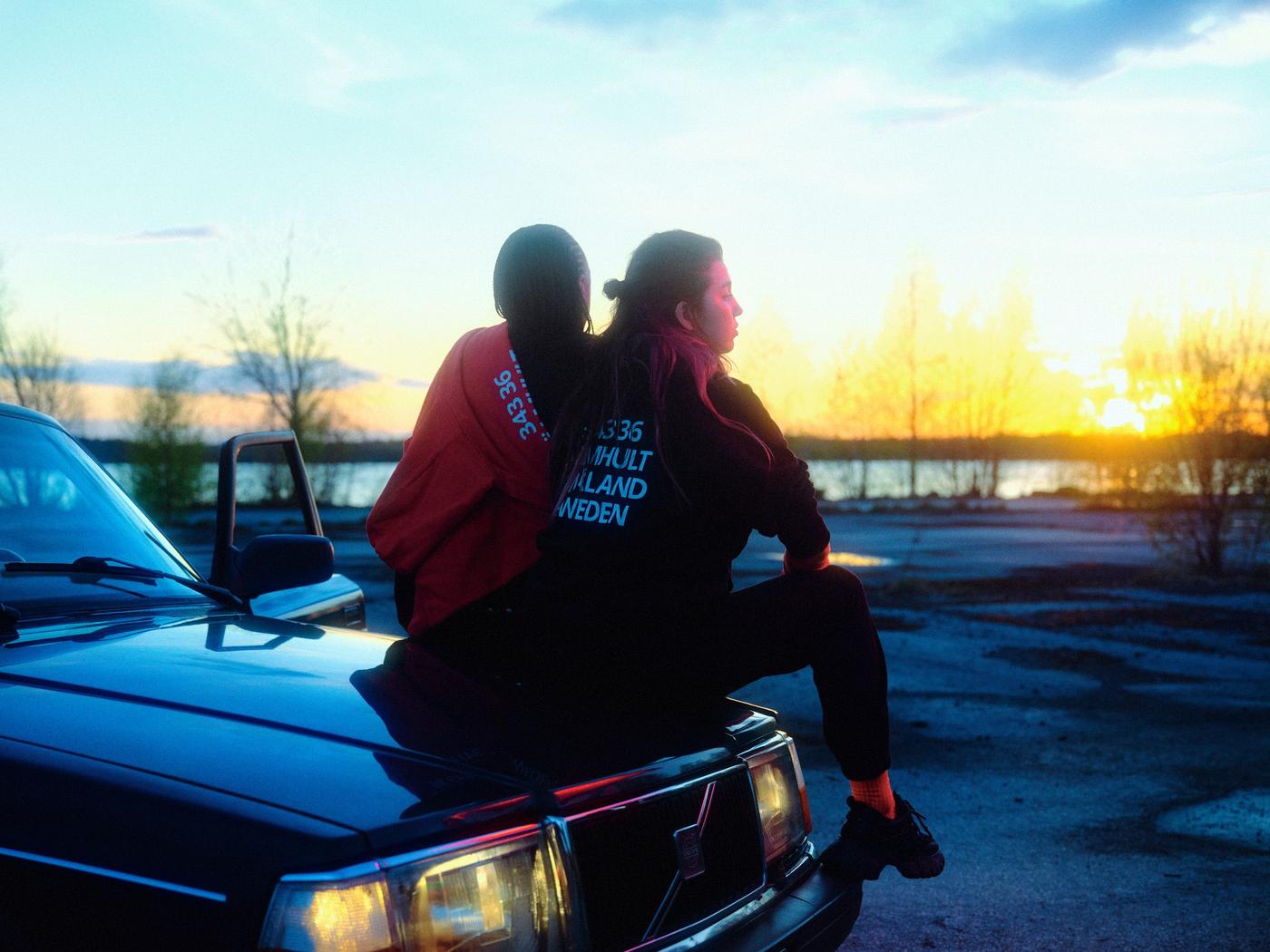 Two people sit on the bonnet of a car in the late afternoon wearing hoodies from the IKEA Museum Älmhult collection.