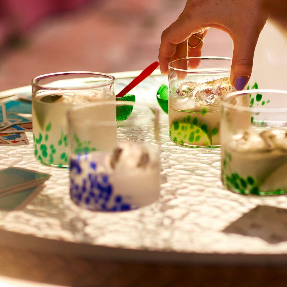 A hand picking up one of four glasses with lemonade, that are standing on a glass table. 