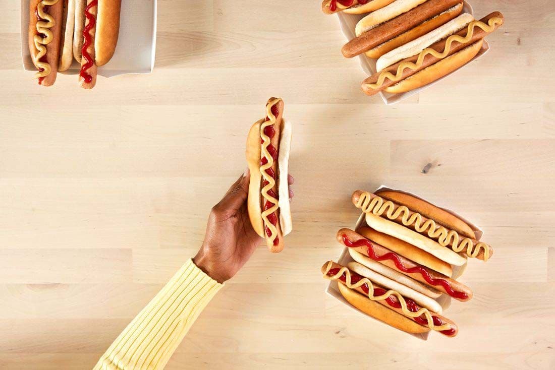 A hand grabs an IKEA plant-based hot dog from a table of hot dogs. The hot dogs are dressed in ketchup and mustard.