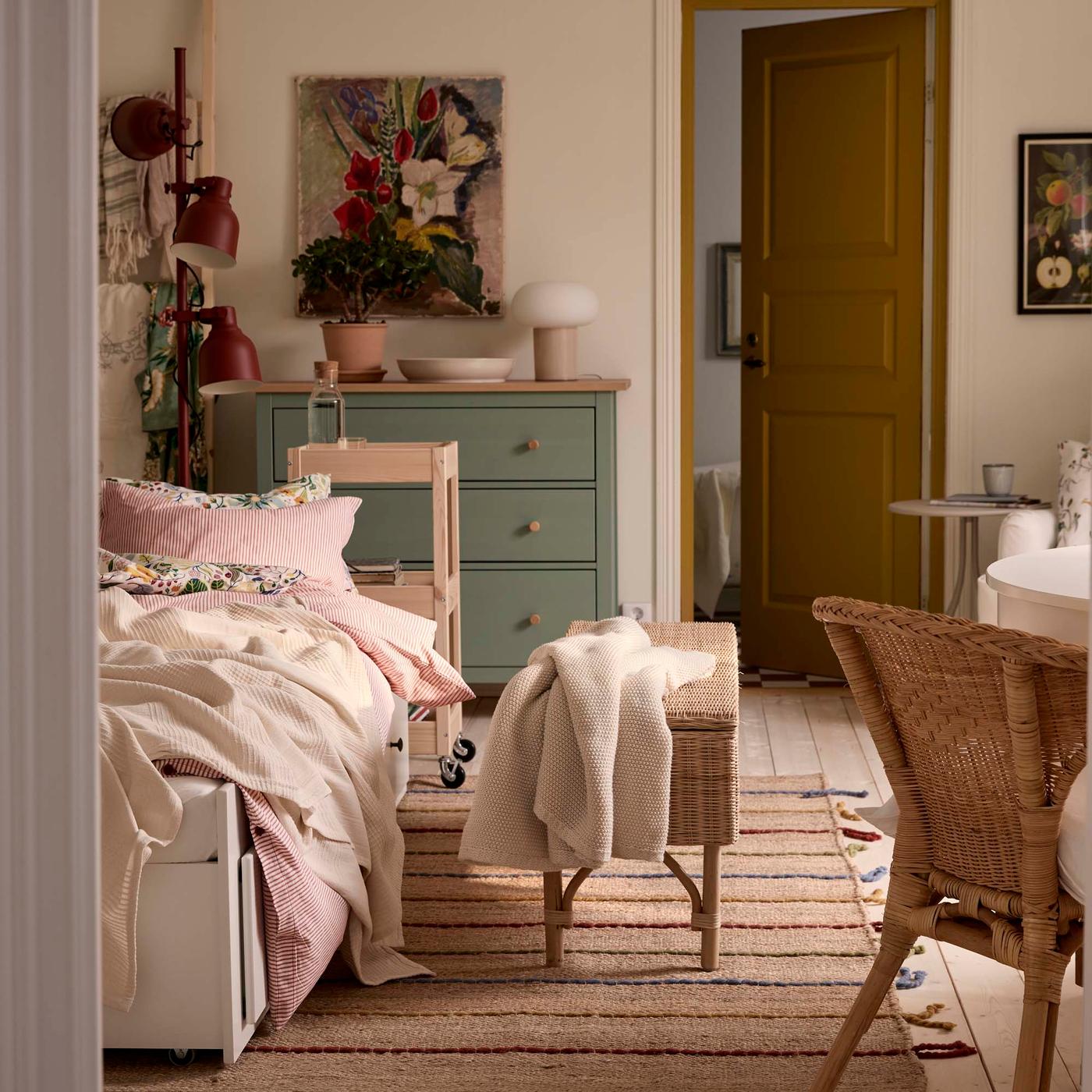 A warm and inviting bedroom with a cozy bed, pastel-coloured bedding, decorative items and a wicker bench with a cream throw laying on top.