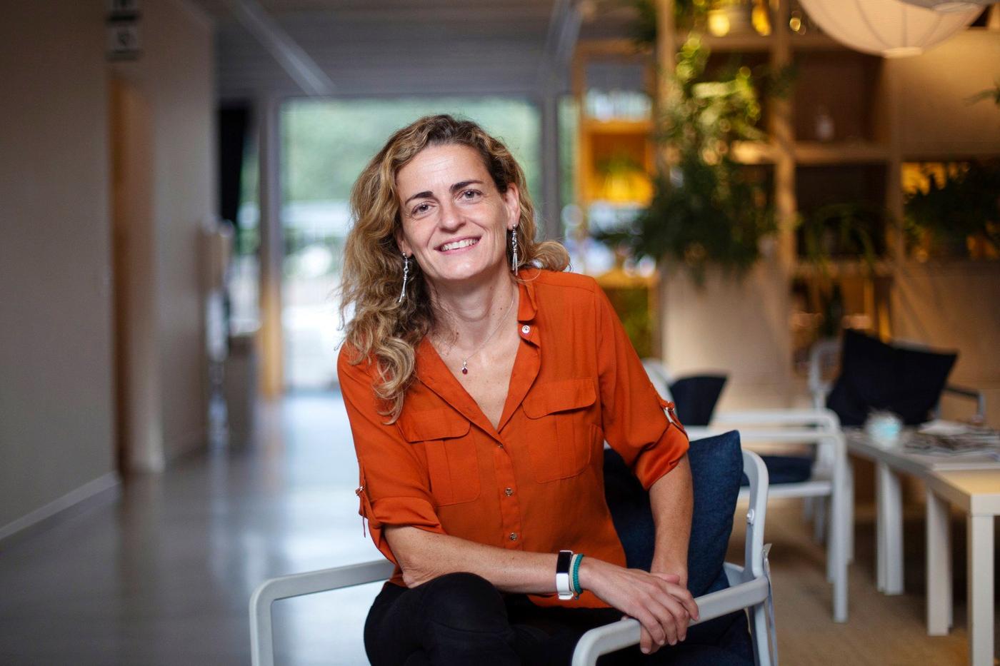 A woman with brown curly hair sitting in a chair in a light office environment.