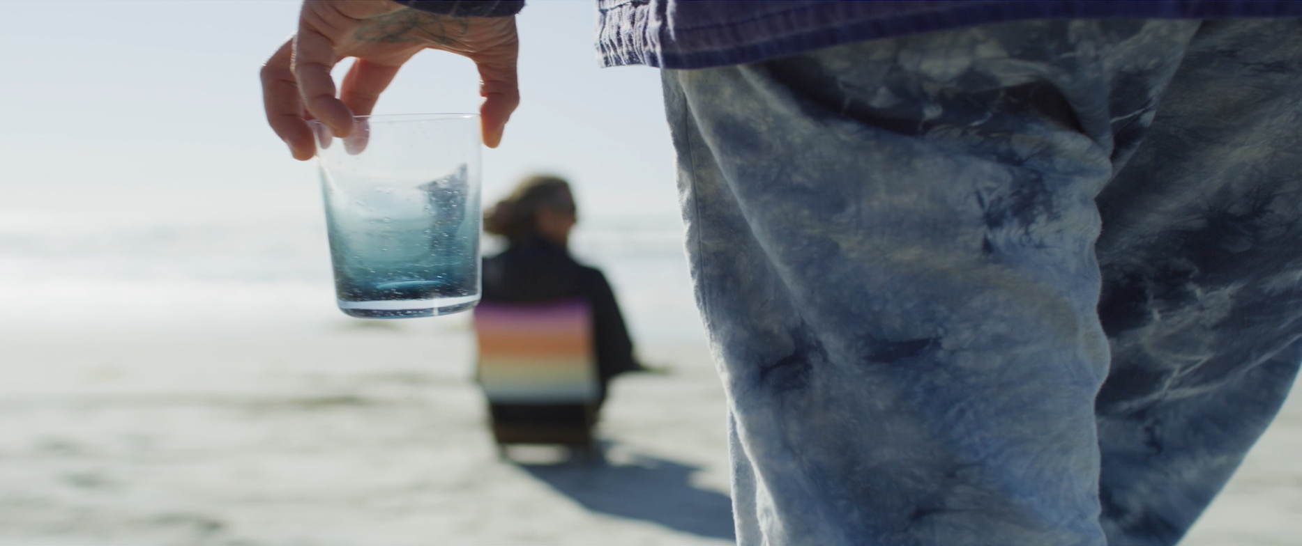 A person carries a blue glass. In the background, another person sits on a chair on the beach.