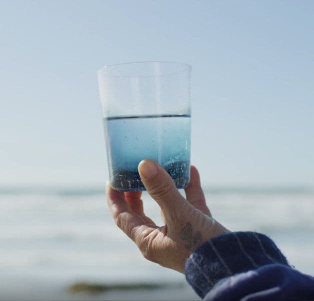 A hand holding up a glass in a pale blue shade from the KÅSEBERGA collection. Blurry ocean waves in the background.