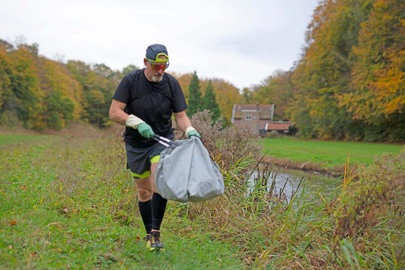A man wearing sports clothes walks along a riverbank. He is picking up rubbish with tongs and putting it in a bag.